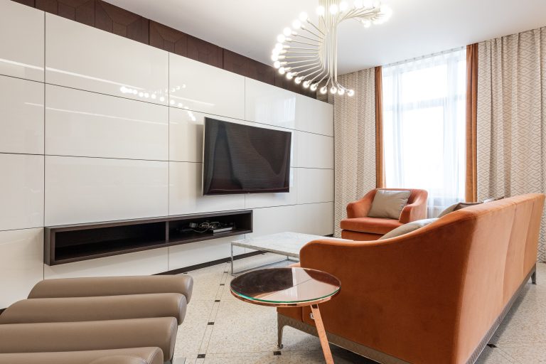 Modern living room with wall-mounted TV, stylish orange sofa set, contemporary chandelier, and large window