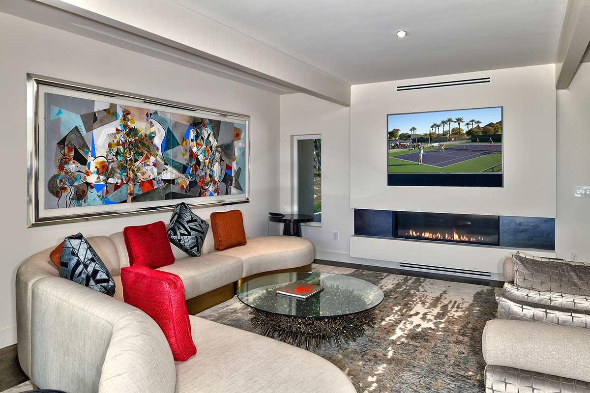Modern living room with a curved sofa, glass coffee table, abstract wall art, gas fireplace, and a wall-mounted TV displaying a tennis match.